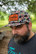 Man wears the black, army green, white and tan camo hat. On the hat a raised, textured bright orange square in the front it says in black text "Franklin Barbecue"