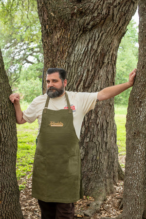 A man stands in the woods. He wears the green apron with stitched on Franklin Barbecue logo in gold