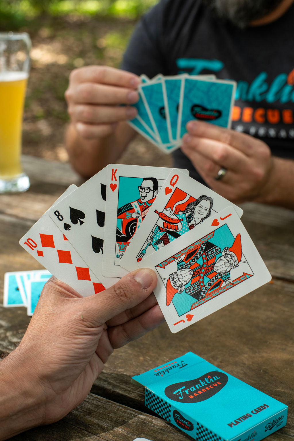 Two people playing cards. We see the hand of one which has a 10 of diamonds, an 8 of spades, The King as portrayed by Aaron Franklin, the Queen as portrayed by Stacy Franklin and the Franklin gnome as the Jack.