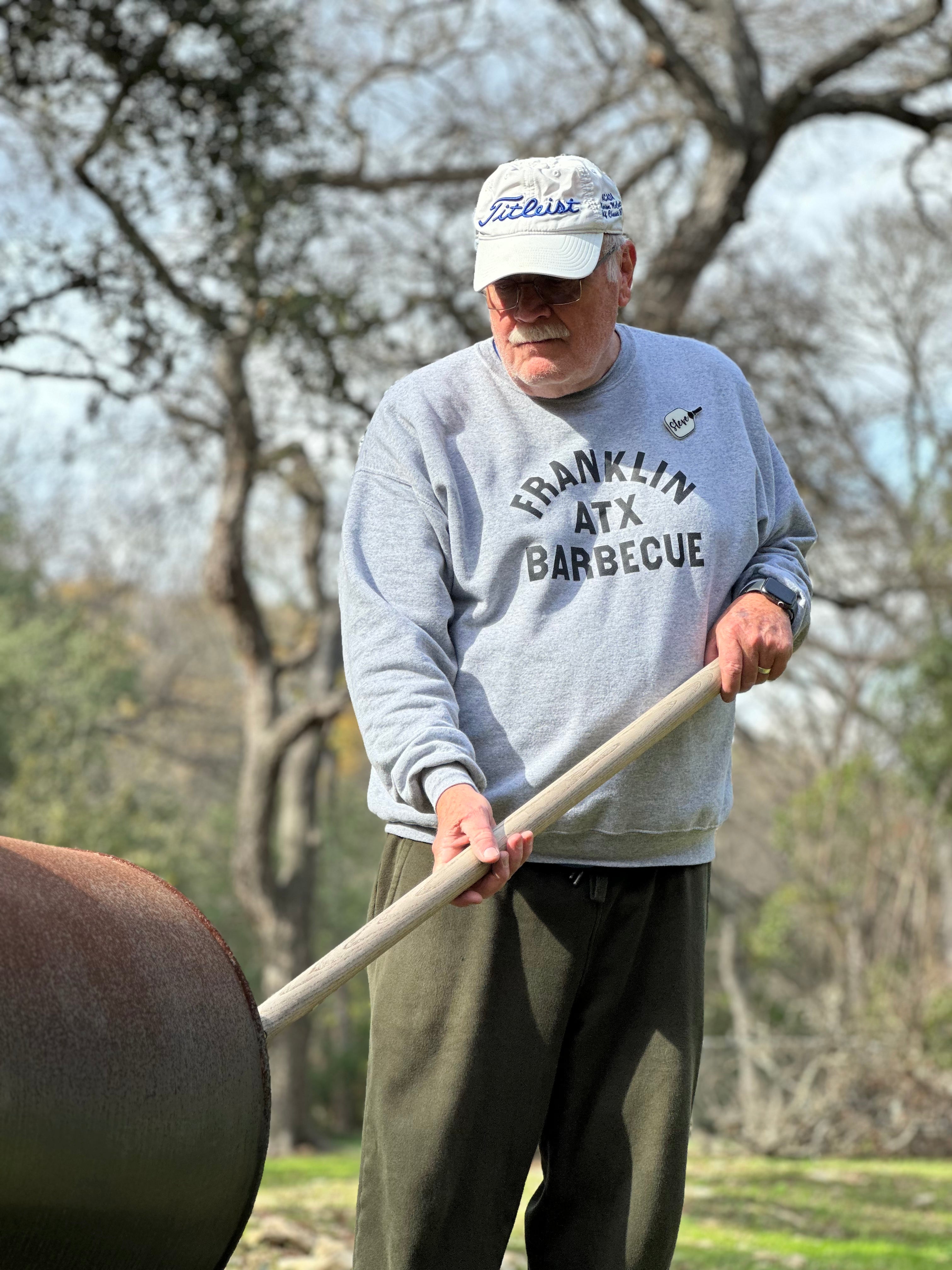 A 71 year old gentlemen is outside and is poking a large wooden stick into a smoker. He wears the grey Franklin ATX Barbecue sweatshirt. He looks calm and comfortable.