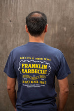 The back of a man in front of a cement wall. He wears the navy blue t-shirt that reads in yellow and white letters: "900 E. 11th St. Austin, TX. Franklin Barbecue. It's served fresh daily until gone. 512-653-1187. Brisket, Pulled pork, Ribs, Turkey, Sausage". Beneath the words is an image of a cloud of smoke over three sausages.
