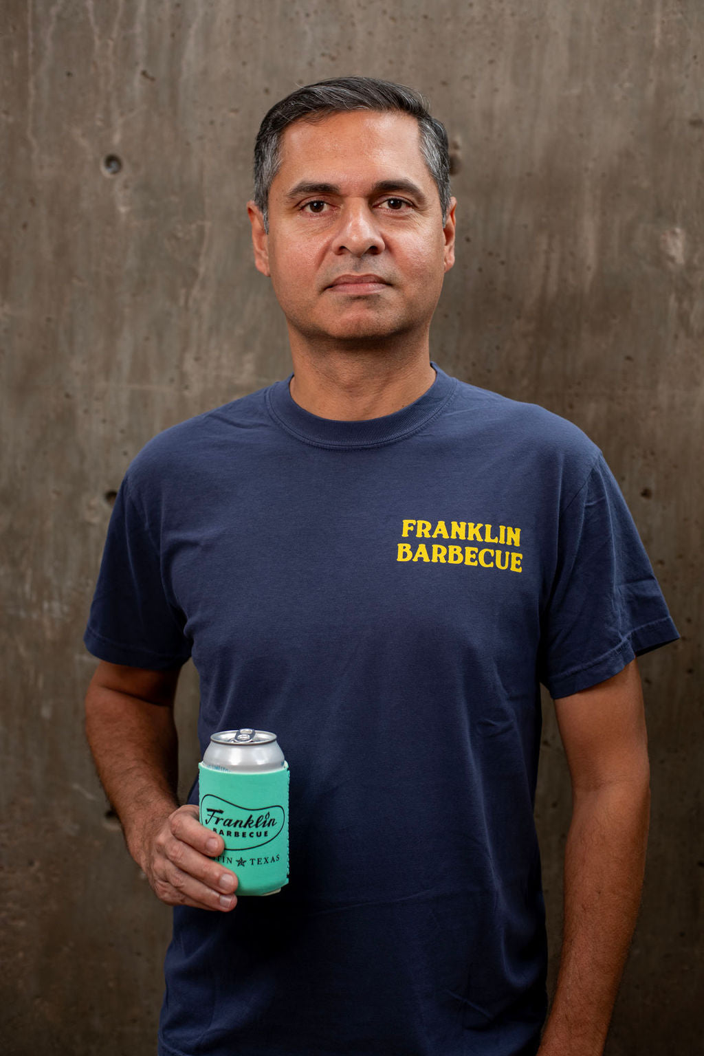 Man in front of a cement wall. He wears the navy blue t-shirt. "Franklin Barbecue" is printed in yellow on the top left side of the shirt. He is also holding a beer covered with our insulated coldie holdie that has our logo on it.