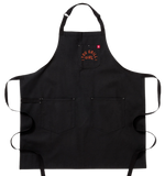 A black apron with two big pockets at the bottom. It has a smaller breast pocket on the left side. On the breast pocket in orange it says "You Grill Girl. Aus Tex".