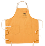 A yellow apron with two big pockets at the bottom. It has a smaller breast pocket on the left side. On the breast pocket in orange it says "You Grill Girl. Aus Tex".