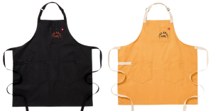 A black apron next to a yellow apron. Both feature two big pockets at the bottom.  Both have a smaller breast pocket on the left side of the apron. On the breast pocket it says "You Grill Girl. Aus Tex". The text is orange on the black apron and black on the yellow apron.