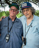Two older men stand in the woods. The one on the right wears the navy hat.