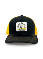 Yellow and black hat that has a white patch on the front with a drawing of a 1950s looking lady holding a spatula. Around the image it says "You Grill Girl". Below that it says "Aus Tex. Les Dames Austin". 