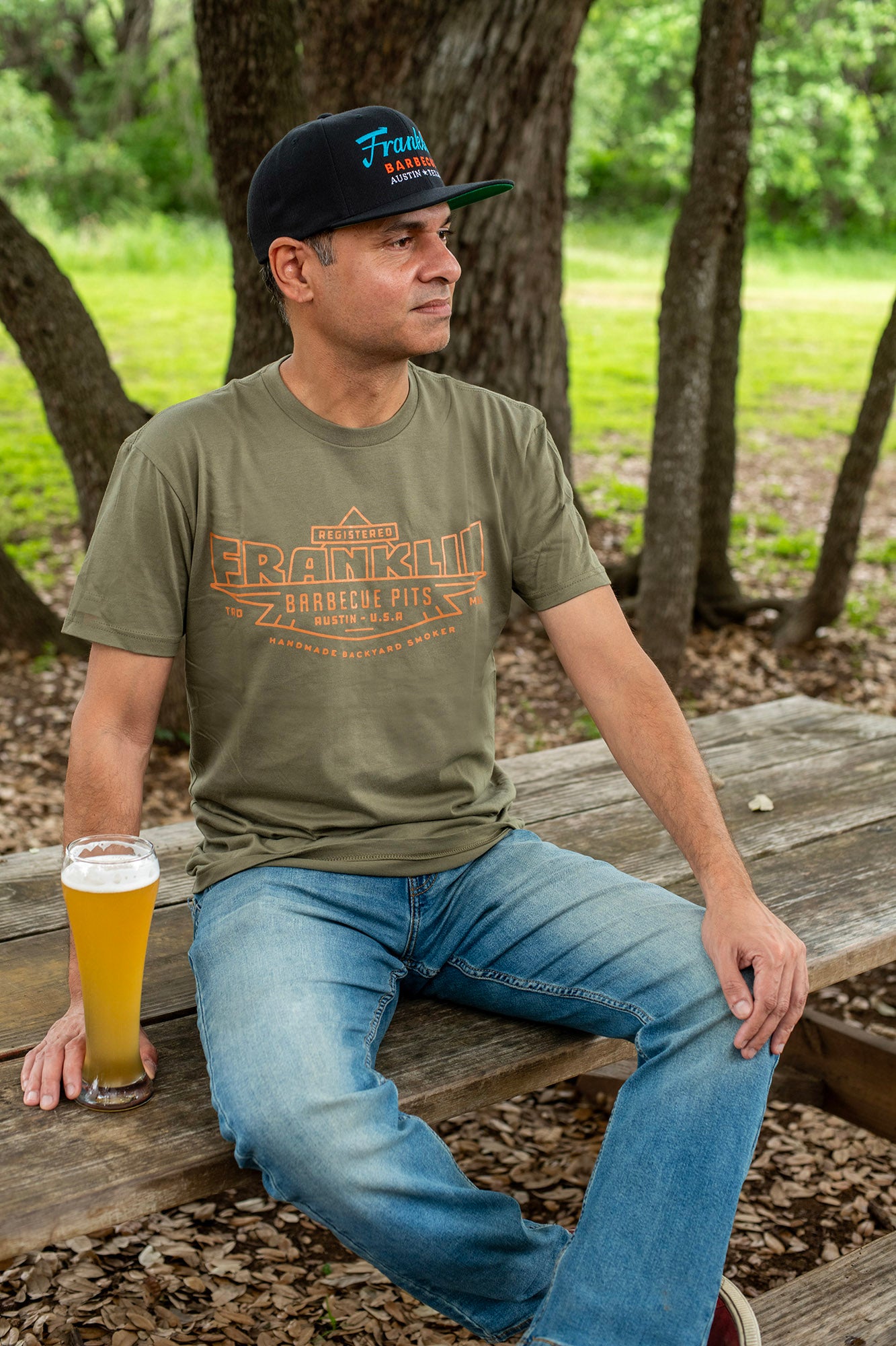 Man sits with a beer, wearing the Army Green t-shirt with Registered Franklin Barbecue Pits logo in orange. He also wears a Black hat with the Franklin logo on it.