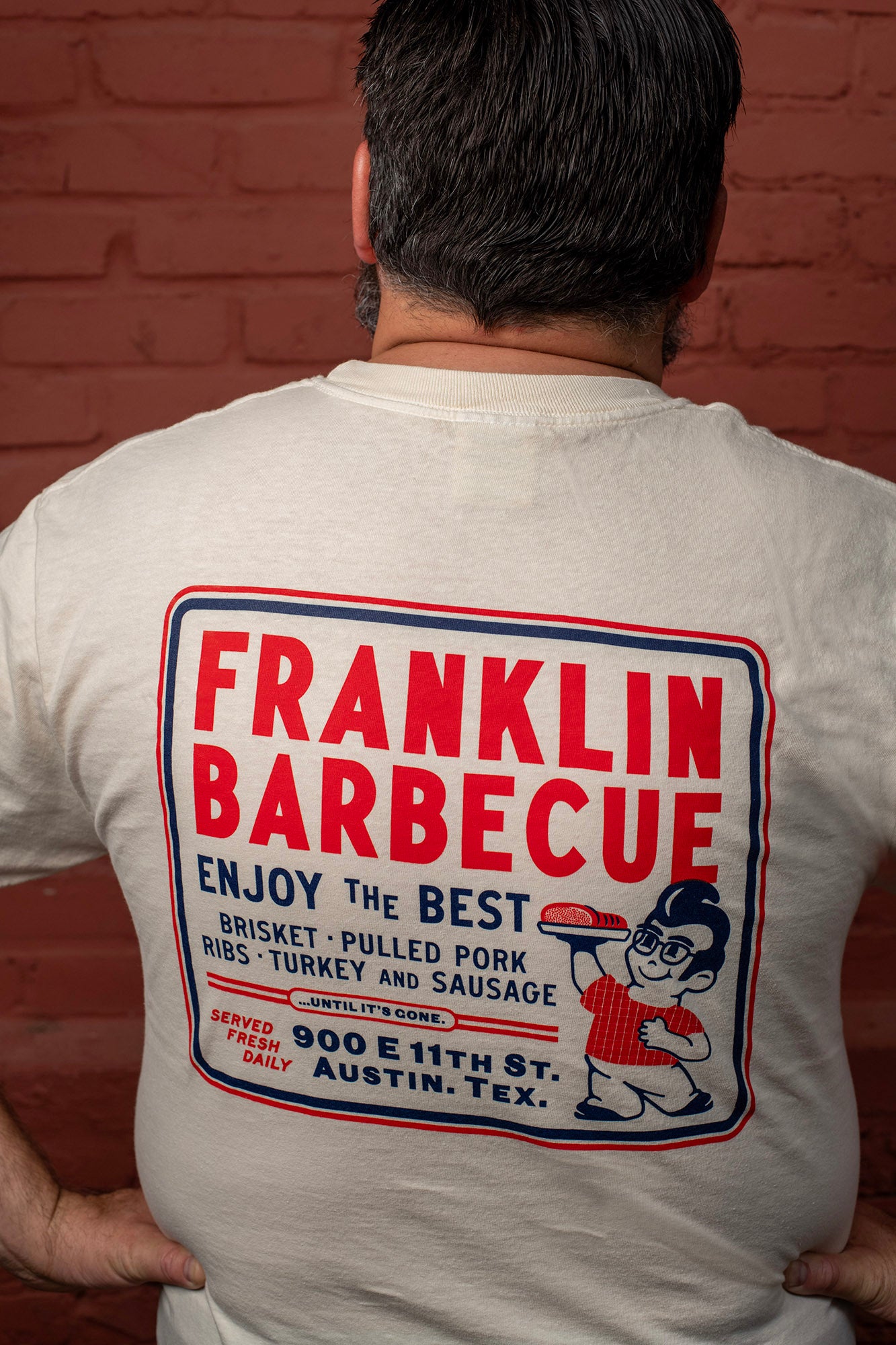 Back of a man against a brick wall. He wears the cream shirt. On it is a big square outlined with blue and red. In the square it says, "Franklin Barbecue. Enjoy the best brisket, pulled pork, ribs, turkey and sausage...until it's gone. Served fresh daily. 900 E. 11th St. Austin, TX." To the right of the font is a drawing of Aaron Franklin holding up a plate of meat.