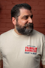 Man with beard looks to the left. He wears the cream t-shirt. On the shirt's top left it says in blue and red letters "Austin, TX. Franklin Barbecue. Enjoy the best."