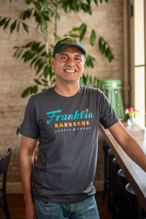 Man modeling Grey t-shirt with Franklin Barbecue text logo in blue and orange. Below that on the shirt is the words "Austin Texas" in white. The man also wears a  Franklin Pits green hat with the Pits logo in Yellow.