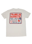 Back side of a cream t-shirt. On it is a big square outlined with blue and red. In the square it says, "Franklin Barbecue. Enjoy the best brisket, pulled pork, ribs, turkey and sausage...until it's gone. Served fresh daily. 900 E. 11th St. Austin, TX." To the right of the font is a drawing of Aaron Franklin holding up a plate of meat. 