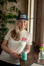 Blonde woman smiles and wears the Navy blue baseball hat with a red band over the brim. A white, black bordered patch on the hat reads in black letters: "Historic East Side Austin Franklin Barbecue Texas. The Best. Served Fresh." She also has a cream colored T-shirt that says "Franklin Barbecue" on the top left side. There is also a coldie holdie, keeping a beer cold that says "Franklin Barbecue".