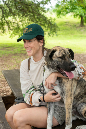 A woman sits with a dog on a picnic bench. She is wearing a dark green hat with logo saying "Registered Franklin Barbecue Pits Austin U.S.A." in yellow