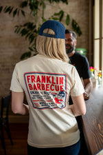 Back of woman at a restaurant. She wears the cream colored shirt. We see the back side of a cream t-shirt. On it is a big square outlined with blue and red. In the square it says, "Franklin Barbecue. Enjoy the best brisket, pulled pork, ribs, turkey and sausage...until it's gone. Served fresh daily. 900 E. 11th St. Austin, TX." To the right of the font is a drawing of Aaron Franklin holding up a plate of meat.