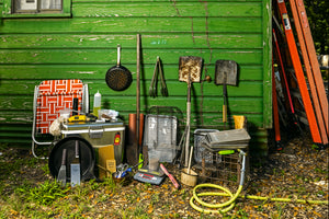 Side of a shed. Leaning against it are tools to aid in cooking barbecue: shovels, cooking oil, a lighter, pans, a scale, etc. 