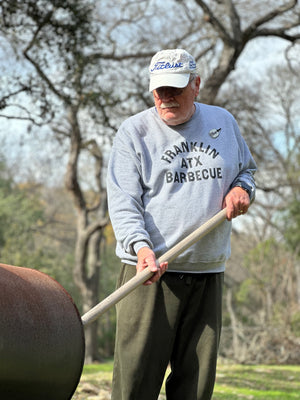 An older gentlemen is outside and is poking a large wooden stick into a smoker. He wears the grey Franklin ATX Barbecue sweatshirt. He looks calm and comfortable.