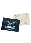 Navy blue postcard with the Franklin Barbecue logo. beneath that is a turquoise drawing of the Franklin restaurant. In white font below that it says, "An Austin original since 2009". We also see the back of the postcard. Franklin's 900 E. 11th St address is listed as the return address. On the top right is a place to put a stamp. On the right side of the card is five lines to write a message. 