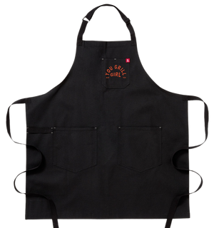 A black apron with two big pockets at the bottom. It has a smaller breast pocket on the left side. On the breast pocket in orange it says "You Grill Girl. Aus Tex".