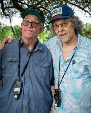 Two older men stand in the woods. The one on the left wears the black hat.