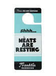 Turquoise blue door hanger. On the top in white text surrounded by black it says "Please do not disturb!" Below that in black text is "Shhh...The Meats are resting". At the very bottom in white text surrounded by black it says "Franklin Barbecue".