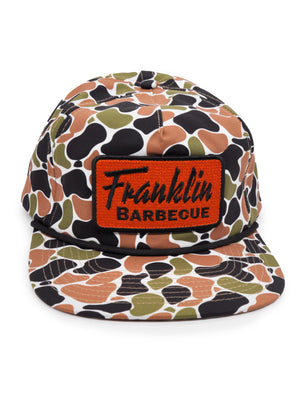 Black, army green, white and tan camo hat. On a raised, textured bright orange square in the front it says in black text "Franklin Barbecue"