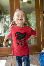Child wearing a Toddler Kids Franklin Barbecue T-shirt in red. T-shirt has the Franklin Barbecue logo with a dancing pig family printed in black.