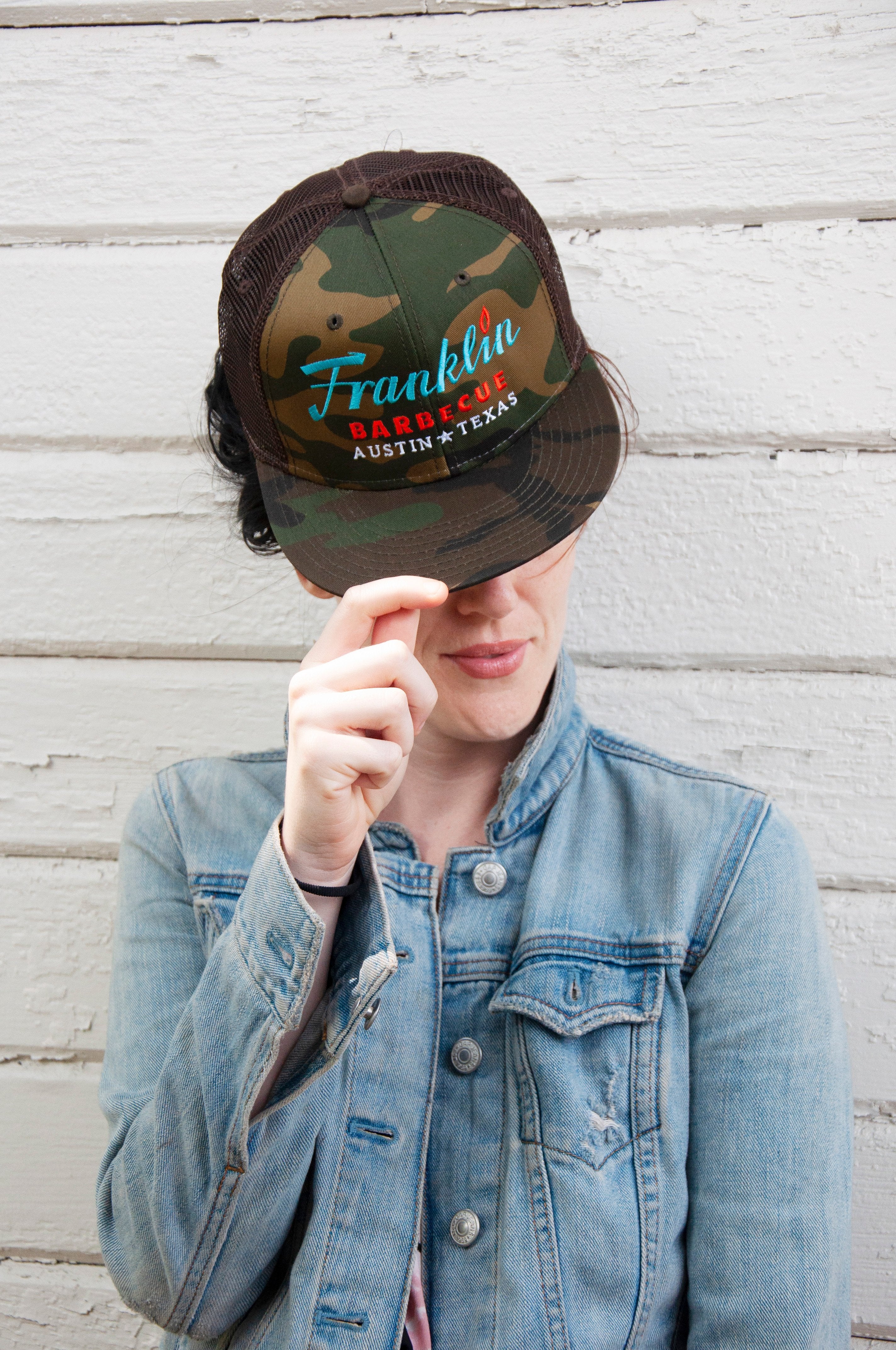 Person modeling Camouflage cap with Franklin Barbecue text logo in blue and orange. Below that is Austin Texas in white.