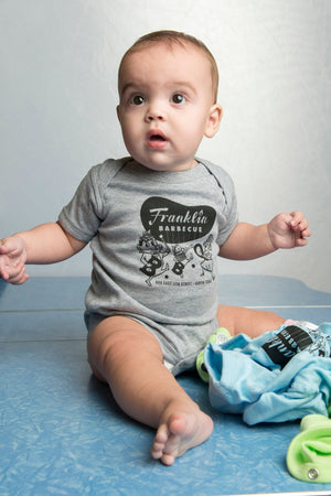 Heather Grey onesie being modeled by baby. Design on onesie is the Franklin Barbecue bean Logo in black and logo of dancing family holding the letters B-B-Q while wearing pig-nose masks also in black. Below the dancing family is the address of Franklin Barbecue also in black. 
