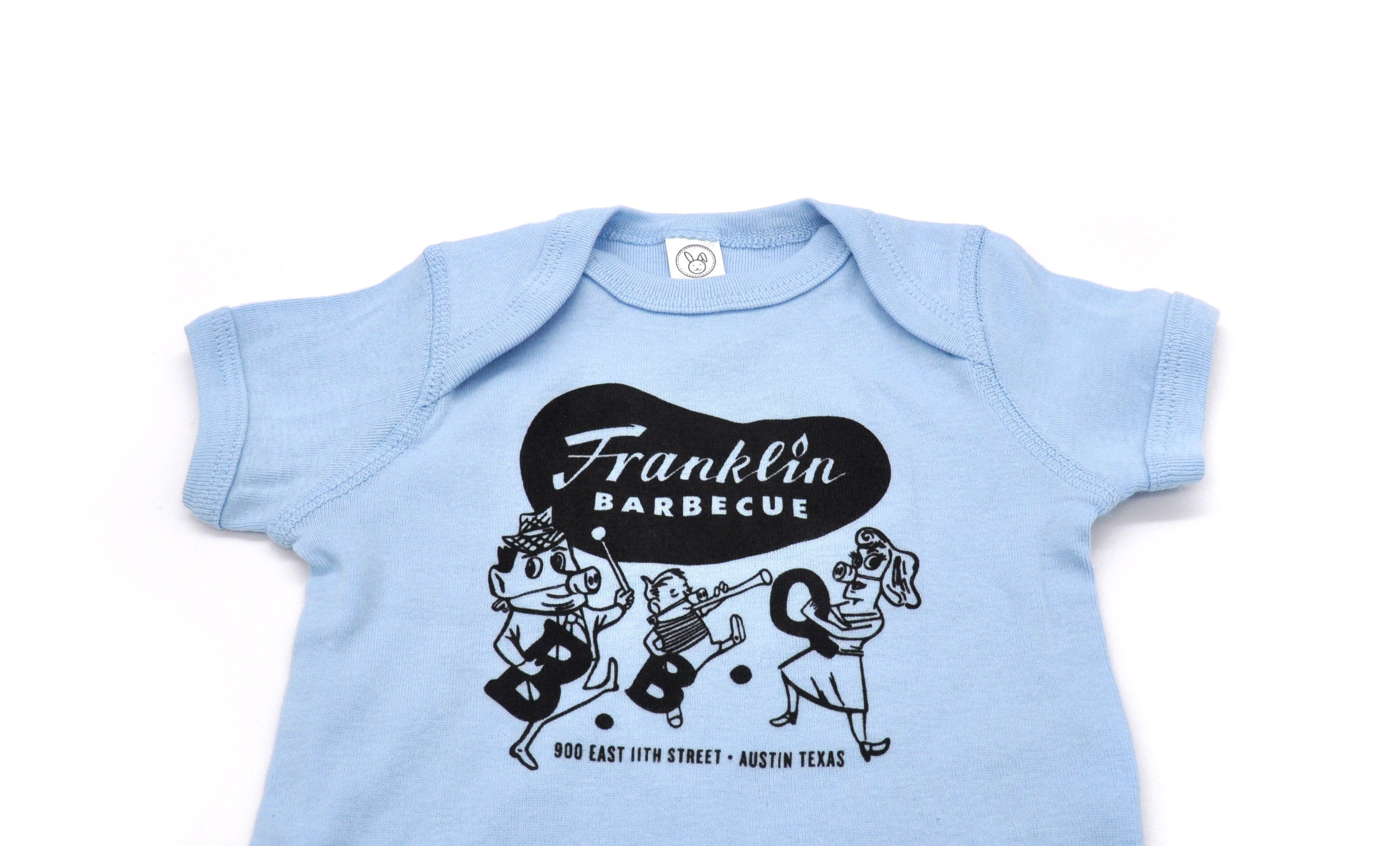 Baby Blue onesie with Franklin Barbecue bean Logo in black and logo of dancing family holding the letters B-B-Q while wearing pig-nose masks also in black. Below the dancing family is the address of Franklin Barbecue also in black.