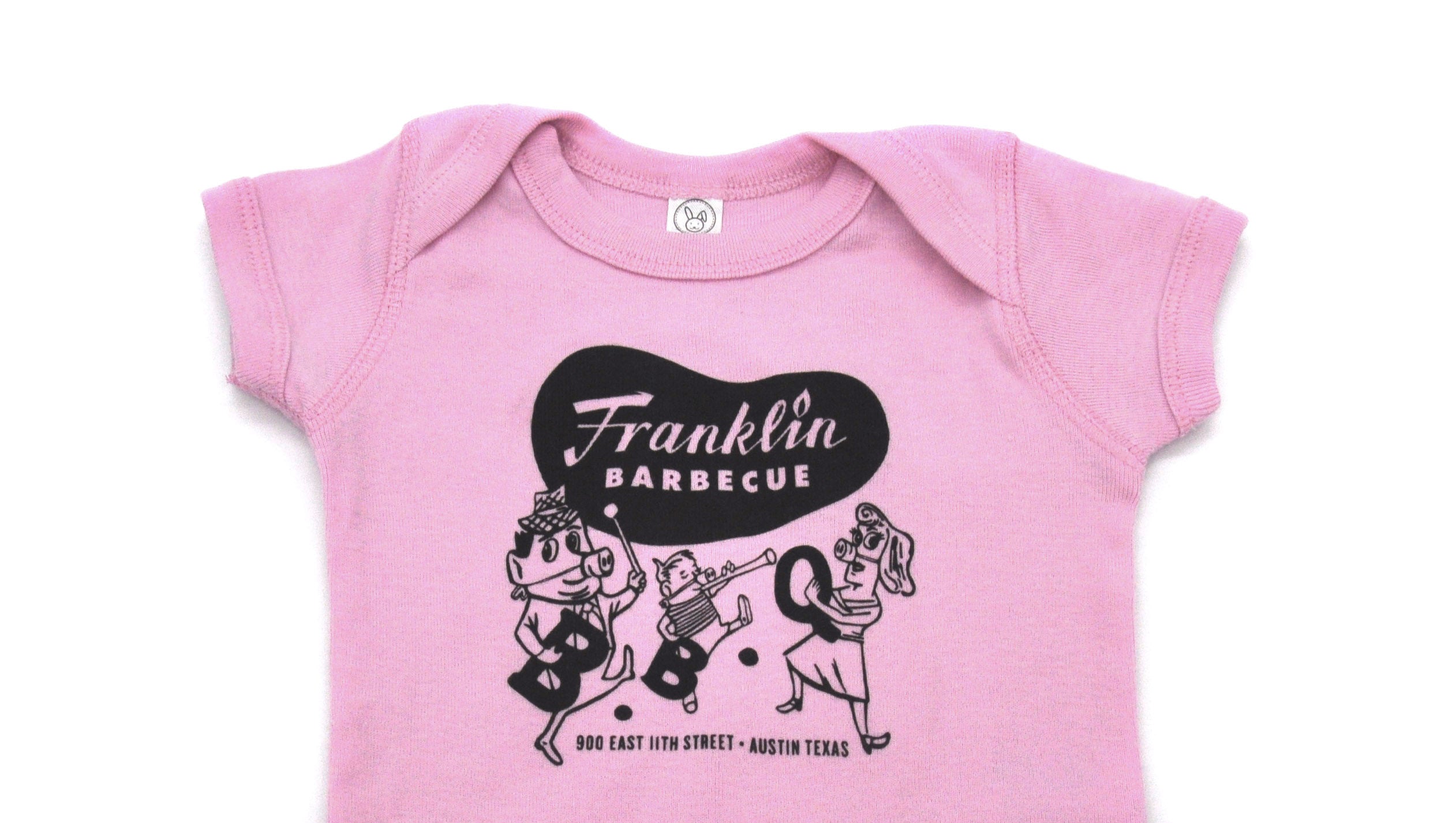 Pink onesie with Franklin Barbecue bean Logo in black and logo of dancing family holding the letters B-B-Q while wearing pig-nose masks also in black. Below the dancing family is the address of Franklin Barbecue also in black.