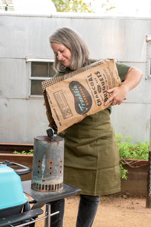 Person pouring charcoal from bag of Franklin Barbecue Charcoal Briquets. Includes Franklin Barbecue bean shaped logo in black with blue and orange lettering