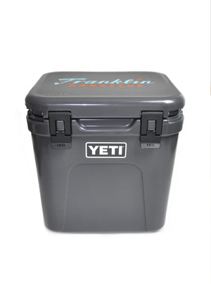 Gray Franklin Yeti Roadie cooler with blue and orange Franklin Barbecue logo on lid and white YETI logo on front face