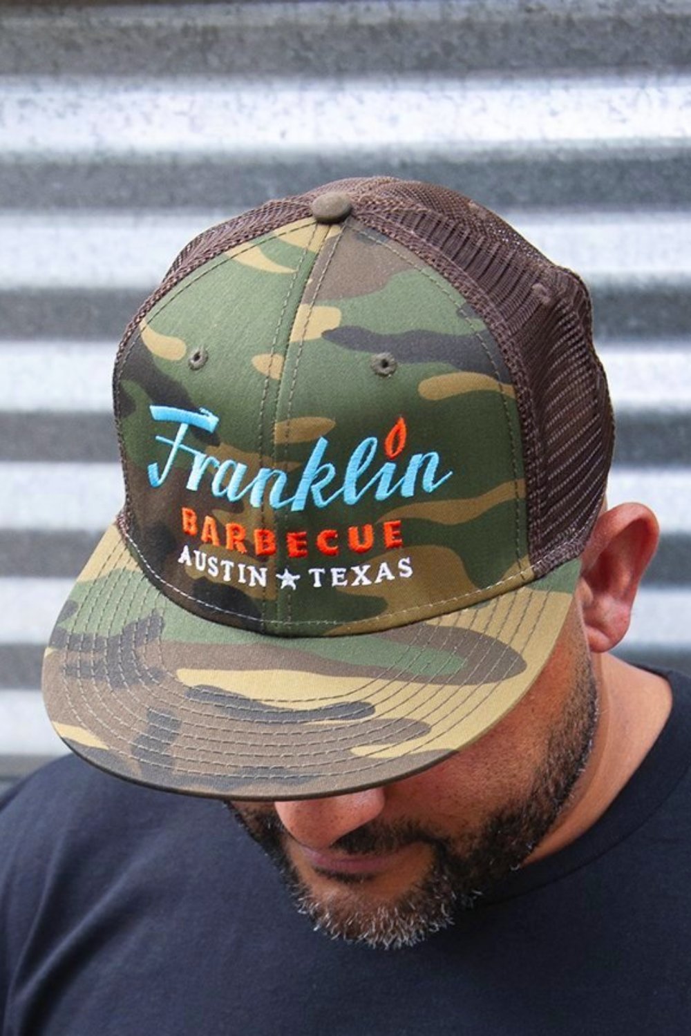 Person modeling Camouflage cap with Franklin Barbecue text logo in blue and orange. Below that is Austin Texas in white.