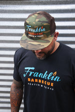 Person modeling Camouflage cap with Franklin Barbecue text logo in blue and orange. He is also modeling Black t-shirt with Franklin Barbecue text logo in blue and orange.