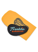 Black patch shaped like Franklin Barbecue bean logo with blue and orange lettering displayed atop coin envelope stamped with black Frankln Barbecue bean logo