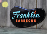 Aluminum tacker sign in the shape of the Franklin Barbecue restaurant sign. Sign is displayed outside on a wooden fence.