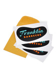 Three Franklin Barbecue restaurant logo stickers laid out atop a yellow coin envelope