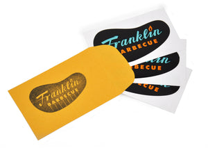 Three Franklin Barbecue restaurant logo stickers laid out atop a yellow coin envelope stamped with Franklin Barbecue restaurant logo