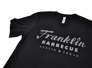 Close up of black short sleeve t-shirt with Franklin Barbecue text logo in dark silver metallic print.