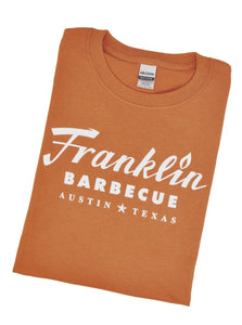 Folded burnt orange shirt.On the short in big white text it says "Franklin Barbecue Austiin Texas".